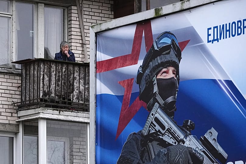 FILE: A woman stands on a balcony in St Petersburg with a billboard calling for a contract for service in the Russian armed forces in the foreground, 4 December 2025