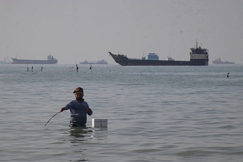 A man stands in the water as bulk carriers, cargo ships, and service vessels line the horizon in the Strait of Hormuz off Bandar Abbas, Iran, Monday, April 27, 2026